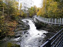 Foto eines Flusses, der steil abfällt. An der rechten Seite befindet sich eine Hängebrücke, links ein größeres Gebäude