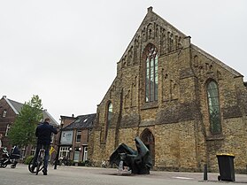Tan stone medieval church with triangular front. The roof is made of glass and there is a modern fountain in the shape of a bat in front. A few pedestrians are milling around.