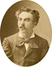 head and shoulders shot of a young man with medium length hair and a substantial moustache