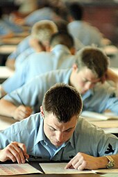Young adults wearing light blue uniforms sit at tables with test papers and pencils