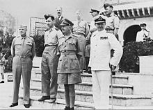 Eight men stand on the steps of a Middle Eastern building. Six wear various uniforms but one wears a business suit.