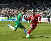 A player dressed in green and white runs with the ball. A player in red and white tried to take the ball. In the background there are stands filled with Hammarby fans.