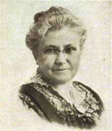 B&W portrait photo of an old woman with her hair in an up-do, wearing a dark blouse, and looking at the viewer.