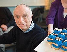 White male in his 70s, wearing a dark blue sweater, to the left of a woman holding a tray of Cookie Monster cupcakes