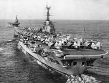 Photograph of an aircraft carrier from behind. Numerous aircraft with their wings folded are sitting on the flight deck. A second aircraft carrier is in the background, leading the first.