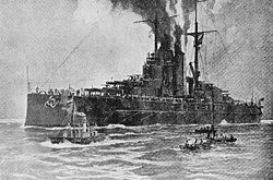 A large battleship sits motionless in the water with smoke coming out of its funnels and three small boats moving beside her in the foreground.