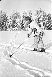 A Finnish soldier on skis, with a fur hat and a tobacco pipe in his mouth, points with a ski pole at the snowy ground where Soviet soldiers have left tracks. The Finnish troops are in pursuit.