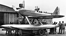 A single-engined monoplane seaplane aircraft is mounted on a wheeled trolley, ten men are standing by the aircraft with one looking into the opened cockpit. The aircraft has 'S1596' painted on the tail.