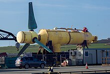 The faded yellow nacelle of a tidal turbine, with three stubby blue blades at the left. Below and in front is a car, portacabin and fencing typical of industrial settings