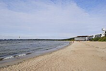 Farbfotografie eines Strandes mit ruhigem Wasser links und feinem Sand rechts. Im linken Hintergrund ist ein Lenkdrachen und im rechten Hintergrund sind weiße Gebäude und Menschen.