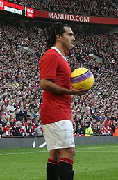 A man wearing a red football shirt, white shorts and black socks. His hair is held back by a hairband. He is holding a yellow football with a purple design.