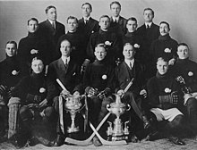 Black and white photo of a hockey team seated behind two large trophies