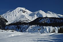 A large snow-covered mountain towering above trees and a snow-covered lake on a clear day