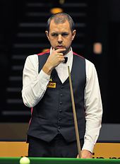Barry Hawkins stands holding his chine with his right hand and a cue in his left hand looking at a table.