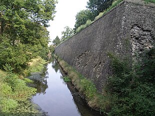 Photo shows an old stone fortress wall about 5 meters tall at right, a wet ditch in the center and trees at left.
