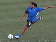 Dwayne De Rosario, wearing a blue jersey, prepares to strike a soccer ball on the ground with his right foot.