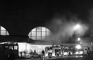 A black and white photograph of King's Cross underground station during the fire with billowing smoke, station lights and fire engines.