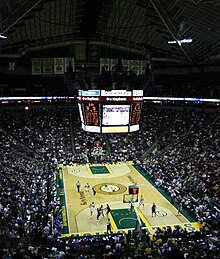 Interior of a sports arena during a game with fans surrounding the basketball court. The overhead video screen has advertisements and a feed of the game displayed alongside statistics.
