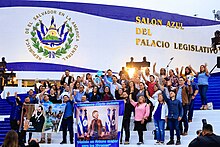 A crowd of people in front of the Legislative Assembly of El Salvador holding banners supporting Nayib Bukele and waving to a camera