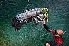 A male diver guides a small aquatic vehicle in a body of water. The vehicle is enclosed in a clear plastic tube and the machinery inside is clearly visible.