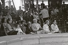 Seitliche Schwarzweißfotografie von Zuschauern in einer Manege. In der vorderen Reihe sitzen von links nach rechts eine Frau, zwei kleine Mädchen, ein Mann und zwei große Mädchen. Sie blicken erstaunt oder lachend nach rechts.
