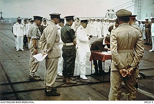 A group of soldiers and sailors parading in various uniforms. In the centre is a small wooden table on which a soldier is signing a document.