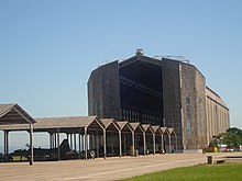 A large tall grey concrete hangar, with the main doors slightly open. In the background, a blue sky. In the foreground are a row of individual aircraft shelters. In one of them, an F-5 is visible in silhouette.