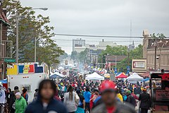 A large street festival in Milwaukee, Wisconsin. Much of the crowd is African American, and cooking smoke can be seen rising from food trucks and stands parallel to the street.