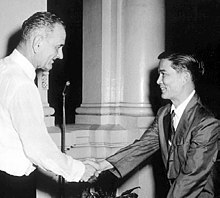 Tall Caucasian man standing in profile at left in a white suit and tie shakes hands with a smaller black-haired Asian man in a white shirt, dark suit and tie.