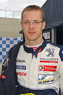 A photograph of a bespectacled Sébastien Bourdais in racing overalls featuring sponsors logos