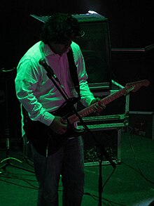 Warren Mendonsa playing an electric guitar onstage, in front of a microphone
