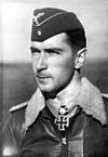 The head and shoulders of a young man, shown in semi-profile. He wears a field cap and a pilot's leather jacket with a fur collar, with an Iron Cross displayed at the front of his shirt collar. His hair is dark and short, his nose is long and straight, and his facial expression is a determined and confident smile; his eyes gaze into the distance.