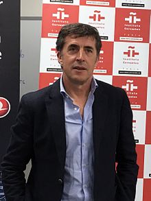 Upper body photo of Delgado in a suit standing in front of an advertising board