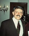 A middle-aged man with a moustache, he is wearing an earth-tone suit and sports a mustache