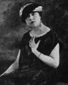 A white woman, seated, wearing a turban-style hat and a loose-fittng black dress with bare arms; she has one hand on her chest