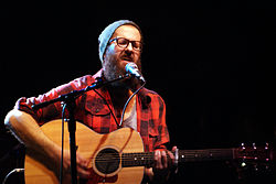 William Fitzsimmons wearing a tuque and a red checkered flannel shirt, playing an acoustic guitar and singing into a microphone onstage