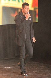 A full body image of Valenciano wearing black pants, shirt, jacket and sneakers while singing into a hand-held microphone