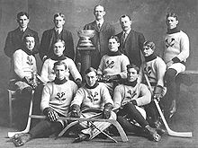 An early ice hockey team poses for a photo. Eight players, all seated around a trophy on a pedestal, are dressed in wool sweaters with a thistle emblem. They wear skates and hold ice hockey sticks. Behind them stand four men in suits.