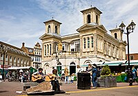 picture of a two story building with turrets and balcony. Market stalls in front and in the foreground is two buskers playing guitars.