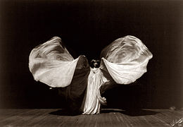 A black and white image of a woman on a stage wearing a dress of long white fabric, which billows out dramatically.