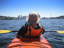 Photo of rear of person wearing orange life preserver sitting in kayak with buildings in far background