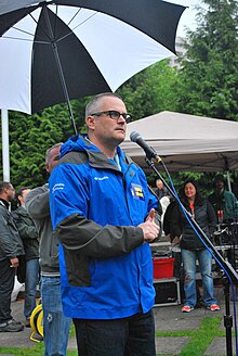 A man behind a microphone stand, wearing glasses and a blue jacket; behind him is a man holding up an umbrella.