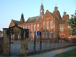 A large red-bricked building with many chimneys, with a small yard and sandstone arch in front