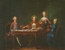 A family seated around a table, playing cards. A young man in red is on the left side and a middle-aged woman in the middle, behind her standing a young woman in pink. On the right side of the table is another young woman in blue.