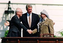 A man in a dark suit on the left shakes the hand of a man in traditional Arab headdress on the right. Another man (Bill Clinton) stands with open arms in the center behind them.