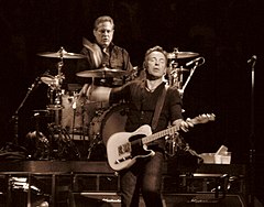 Middle-aged man with glasses wearing a dark shirt sits behind a drum kit on a riser with his right hand and stick about to hit a snare drum; he his looking directly at another middle-aged man in front of and slightly to the left of him, dressed in dark clothes with an electric guitar strapped to him, left hand on the frets, right hand flying away after playing a chord, eyes shut in an expression of conveying musical intensity of some kind