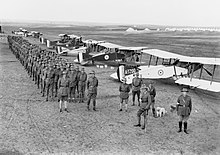 Black and white photograph of a large group of men dressed in military uniform standing in close formation next to a row of biplane aircraft