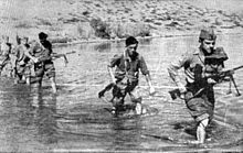 a black and white photograph of a line of males in uniform carrying weapons wading through a shallow river