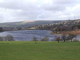 A large reservoir surrounded by fields, hills and trees, with a village on the background hill