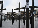 A photograph of a large number of black crosses with photographs on them standing on white-speckled stone all under a blue sky with white clouds